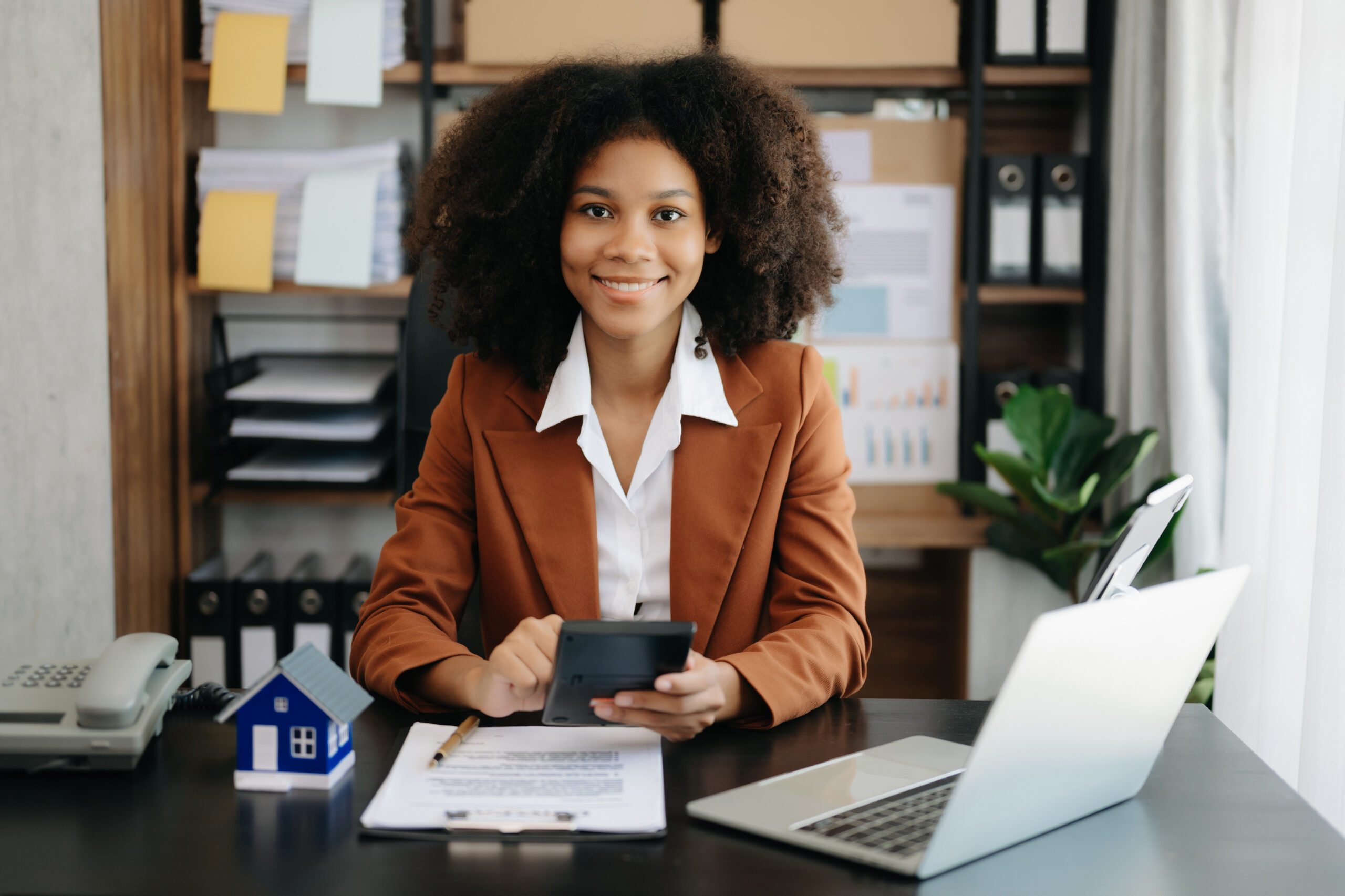 Young real estate agent worker working with laptop and tablet at table in office and small house beside it. Eine Versicherungsexpertin berechnet Beiträge